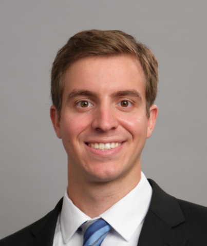 headshot of Quinn with a white collared shirt and a black blazer with a grey background 