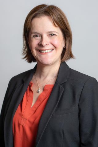 Headshot of R Ratner with gray background. Ratner is wearing a Black blazer and red blouse
